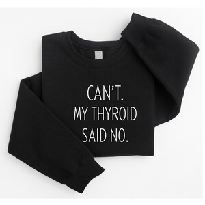 May include: Black sweatshirt with the text "CAN'T. MY THYROID SAID NO." in white. The sweatshirt is folded, with the sleeves and neckline visible. The background is plain white.