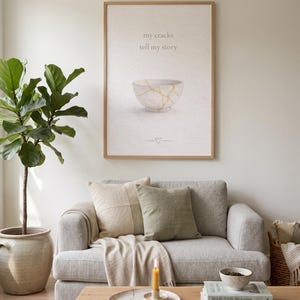 Puede incluir: Una impresión enmarcada con el texto "my cracks tell my story" encima de una ilustración de un cuenco blanco con detalles dorados. Un sofá gris con cojines y una mesa de centro de madera con libros y una vela.