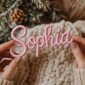 May include: A pink embroidered patch spelling "Sophia" in a cursive script. The patch is displayed against a cream-coloured knitted jumper. The background features a pine cone and fir branches.