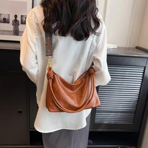 May include: A brown leather shoulder bag with a knotted handle and a patterned strap. The bag is a warm shade of brown and has a simple, elegant design. The strap is a mix of blue, brown, and gold.