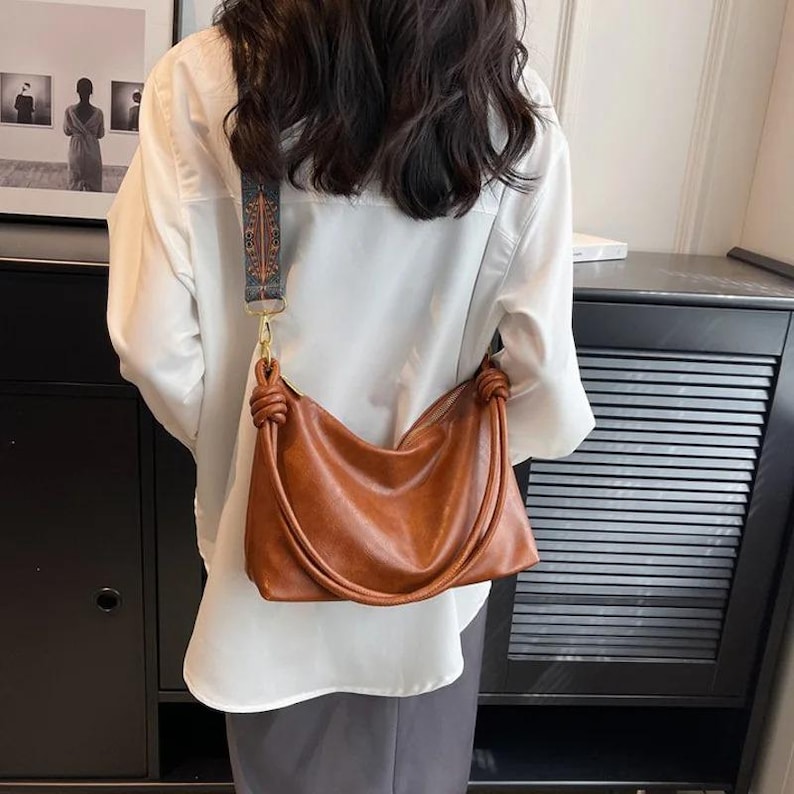 May include: A brown leather shoulder bag with a knotted handle and a patterned strap. The bag is a warm shade of brown and has a simple, elegant design. The strap is a mix of blue, brown, and gold.