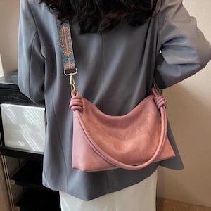 May include: A dusty rose shoulder bag with a knotted handle and a patterned strap. The bag has a curved shape and a gold-colored clasp. The strap features a design with blue, orange, and brown accents.