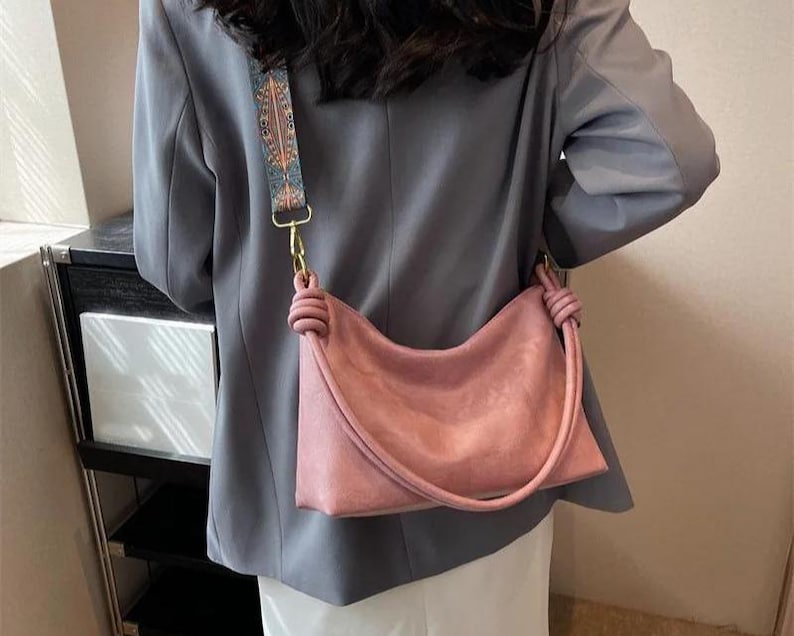 May include: A dusty rose shoulder bag with a knotted handle and a patterned strap. The bag has a curved shape and a gold-colored clasp. The strap features a design with blue, orange, and brown accents.