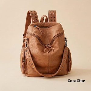 May include: A tan leather backpack featuring a textured surface, multiple zippered compartments, and adjustable shoulder straps adorned with small metal studs. The bag includes a top handle and side pockets, with the text ZoraZinc visible at the bottom.
