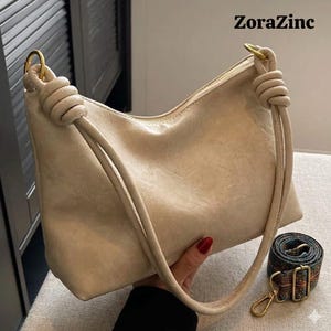 May include: A beige shoulder bag with a knotted handle and a gold-tone metal ring. The bag has a zipper closure and a detachable patterned strap. The text "ZoraZinc" is visible in the upper right corner.