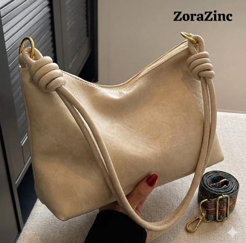 May include: A beige shoulder bag with a knotted handle and a gold-tone metal ring. The bag has a zipper closure and a detachable patterned strap. The text "ZoraZinc" is visible in the upper right corner.