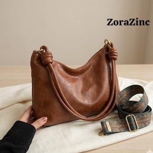May include: A brown leather shoulder bag with knotted handles and a patterned strap. The bag has a curved shape and gold-colored hardware. The text "ZoraZinc" is visible in the upper right corner.