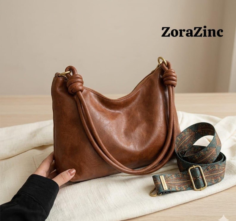 May include: A brown leather shoulder bag with knotted handles and a patterned strap. The bag has a curved shape and gold-colored hardware. The text "ZoraZinc" is visible in the upper right corner.