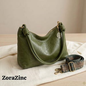 May include: Olive green shoulder bag with a knotted handle and a detachable patterned strap. The bag features a gold-tone zipper and a leaf charm. The text "ZoraZinc" is printed on the bottom left.
