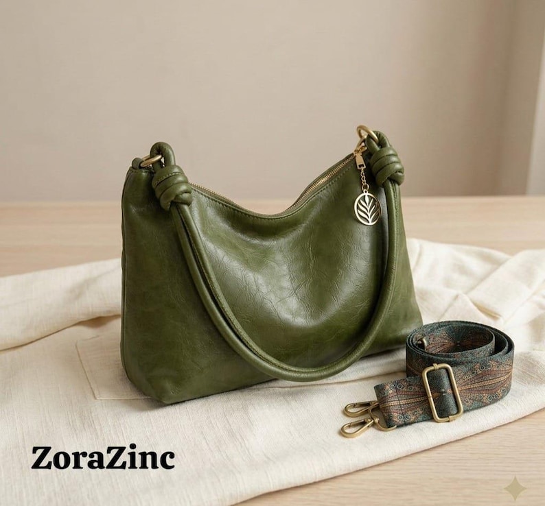 May include: Olive green shoulder bag with a knotted handle and a detachable patterned strap. The bag features a gold-tone zipper and a leaf charm. The text "ZoraZinc" is printed on the bottom left.