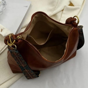 May include: A brown leather handbag with an open zipper, revealing interior pockets. The bag features a patterned strap in shades of teal, brown, and orange. Gold-colored hardware accents the bag's design, adding a touch of elegance.