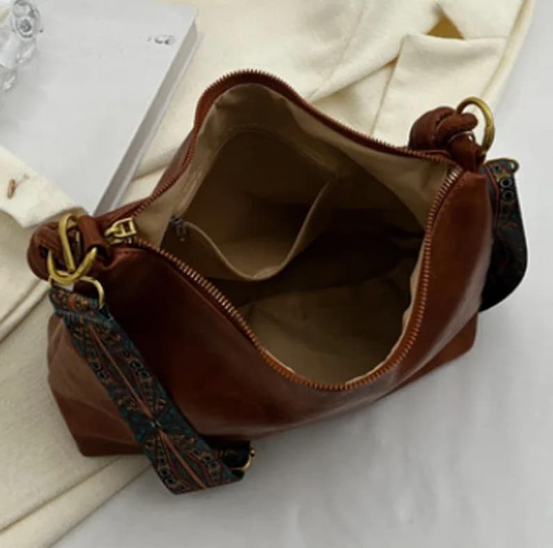 May include: A brown leather handbag with an open zipper, revealing interior pockets. The bag features a patterned strap in shades of teal, brown, and orange. Gold-colored hardware accents the bag's design, adding a touch of elegance.