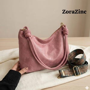 May include: A dusty rose-colored handbag with a knotted handle and a detachable patterned strap. The bag has a curved shape and a zipper closure. The text "ZoraZinc" is visible in the upper right corner.