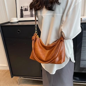 May include: A brown leather shoulder bag with a knotted handle and a gold zipper. The bag is worn over the shoulder and has a long strap. The bag is a light brown color and appears to be made of soft leather.