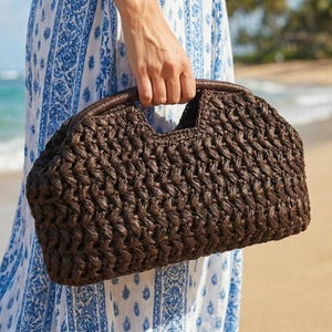 May include: A dark brown woven handbag with a curved top and a handle. The bag has a textured pattern and a leather handle. The bag is held by a person on a beach.