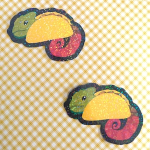 May include: Two stickers featuring a cartoon chameleon with a taco shell body. The chameleon is green with a yellow taco and a red, glittery tail. The stickers are on a yellow and white checkered background.