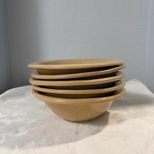 Może przedstawiać: Stos pięciu pasujących do siebie misek ceramicznych w kolorze opalenizny o gładkim, błyszczącym wykończeniu i szerokim, zaokrąglonym brzegu. Miski są jednolite pod względem koloru i kształtu, charakteryzują się prostym, funkcjonalnym designem.