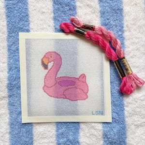 May include: A pink flamingo cross-stitch design on white canvas, with pink embroidery floss. The flamingo is in a resting pose, with a yellow beak and a purple wing. The canvas is on a blue and white striped towel.