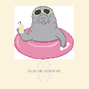Puede incluir: Ilustración pixelada de un manatí gris con gafas de sol flotando en una boya rosa. El manatí sostiene un vaso con una bebida amarilla y una pajita roja. El texto "LUCKY GIRL NEEDLEPOINT" está debajo de la imagen.