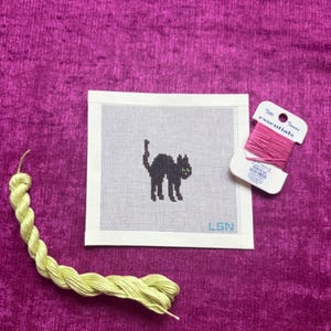 May include: A square needlepoint canvas featuring a black cat silhouette with green eyes and the letters LGN in the bottom right corner. A skein of light green embroidery floss and a spool of pink thread labeled Tilli Tomas essentials are included.