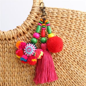 Op de afbeelding: Een decoratieve tashanger met een bronskleurige metalen karabijnhaak, veelkleurige houten kralen, een grote rode pompon, een felroze kwastje en een rond geborduurd stoffen accent met levendige bloemmotieven in diverse kleuren.