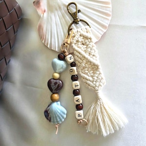 May include: A handcrafted keychain with a gold-tone clasp, incorporating a cream macrame element and a tassel. The keychain includes wooden beads spelling "PATTON", alongside heart-shaped and shell-shaped charms in blue, brown, and cream colours.
