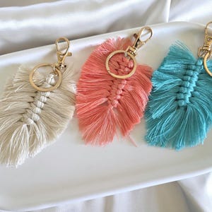 May include: Three macrame feather keychains in cream, coral, and teal. Each keychain features a gold-toned metal lobster clasp and a circular ring attached to a knotted cotton cord design that mimics the shape and texture of a natural feather.