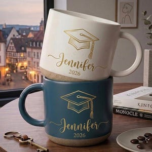 May include: A cream-coloured ceramic mug stacked on top of a dark blue ceramic mug. Both mugs feature a gold-coloured graphic of a graduation cap, the name Jennifer in cursive script, and the year 2026 printed in a simple serif font.