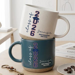 May include: Two ceramic mugs stacked vertically. The top mug is cream with speckles, and the bottom mug is dark teal. Both feature the text "class of 2026" with a graduation cap icon, the name "Jennifer," and "NORTH WASHINGTON HIGH SCHOOL."