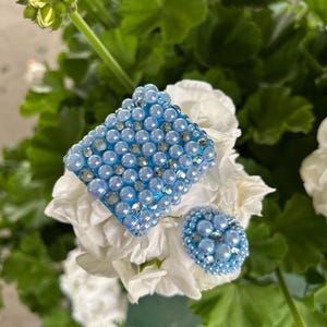May include: A square-shaped accessory and a smaller oval-shaped accessory, both covered in light blue iridescent beads and small clear crystals, resting on white flower petals with green leaves in the background.