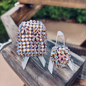 May include: A rectangular case and a circular case, both covered in iridescent, multi-coloured rhinestones. Each item features small, light pink bow embellishments integrated among the sparkling crystals, displayed on clear plastic stands.