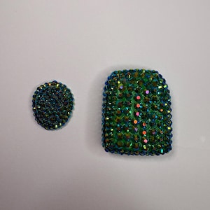May include: Two decorative items, a round button and a rectangular shape, both covered in iridescent green and blue crystals. The crystals catch the light, creating a sparkling effect. The items are likely for embellishment or decoration.