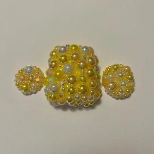 Op de afbeelding: Een set van drie gele en parelachtige, met kralen bezette accessoires. Het centrale stuk is rechthoekig, geflankeerd door twee kleinere, ronde stukken. De kralen variëren in grootte en hebben een glanzende, iriserende kwaliteit.
