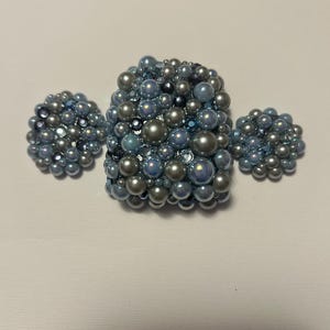 Puede incluir: Un conjunto de joyas que incluye un broche cuadrado y dos pendientes redondos, adornados con perlas iridiscentes azules y plateadas. Las perlas, de diferentes tamaños, crean un efecto texturizado y agrupado. El conjunto está diseñado para brillar.
