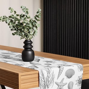 Op de afbeelding: Een zwarte vaas met een groene plant staat op een houten tafel. Een witte tafelloper met een grijswaarden schelpenpatroon is zichtbaar. De tafelloper heeft verschillende schelpen, zeesterren en zeewier ontwerpen.