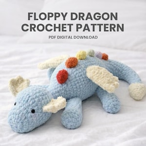 May include: A light blue crocheted dragon toy with cream-coloured wings and accents. The dragon has colourful pom-poms on its back and the text "Floppy Dragon Crochet Pattern" is displayed above. PDF digital download.