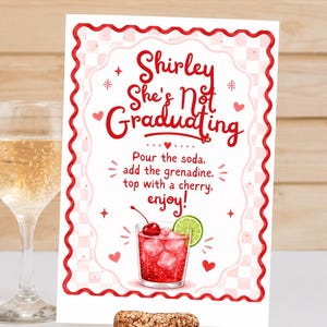 May include: A greeting card with a red and white border and the text "Shirley She's Not Graduating." The card features a watercolor illustration of a cocktail with a cherry and lime. A cork sits in front of the card.