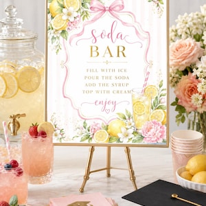 May include: A decorative sign for a soda bar, featuring the text "Soda Bar" in elegant script. The sign is adorned with floral and lemon illustrations, and sits on a gold easel. Other items include drinks, fruit, and a graduation cap.