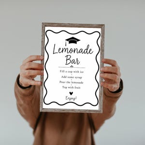 May include: A framed sign with the words "Lemonade Bar" in a decorative font. The sign includes instructions: "Fill a cup with ice, Add some syrup, Pour the lemonade, Top with fruit, Enjoy!" The frame is a light brown colour.