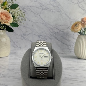 May include: A silver-tone Seiko automatic wristwatch with a cream-colored dial, featuring Arabic numerals at the 6, 9, and 12 o'clock positions, a day-date window at the 3 o'clock position, and a stainless steel link bracelet with a fold-over clasp.