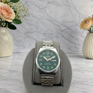 May include: A silver-tone Seiko automatic wristwatch with a round green dial, luminous circular hour markers, a day-date window at the three o'clock position, and a stainless steel link bracelet. The dial features the text Seiko, Sports, and Diver's 200m.