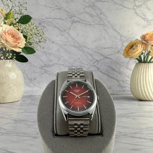 May include: A stainless steel wristwatch with a red dial and a silver-tone metal band. The watch is displayed on a gray stand. The watch face displays the day and date. The background includes a marble-patterned surface and vases with flowers.