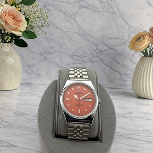 May include: A silver-tone wristwatch with a salmon-colored dial, featuring a stainless steel link bracelet, luminous hour markers, silver hands, and a day-date window at the three o'clock position with the text Seiko printed on the watch face.