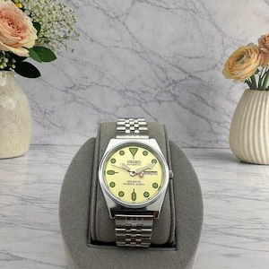 May include: A silver-tone Seiko automatic wristwatch with a pale yellow dial, round green hour markers, and a stainless steel link bracelet. The watch face displays the text Seiko Automatic Sports Diver's 200m and a day-date window at the 3 o'clock position.