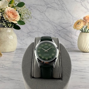 Puede incluir: Un reloj Oris con esfera verde y correa de cuero verde oscuro. El reloj tiene detalles plateados y números blancos. El reloj está etiquetado como "17 JEWELS SHOCK PROOF". Se muestra sobre un soporte gris.