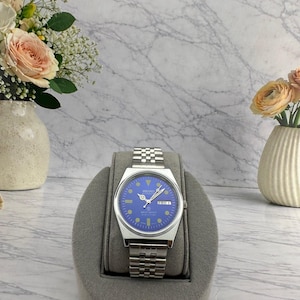 May include: A stainless steel wristwatch with a vibrant blue dial, silver-tone hands, and circular hour markers. The watch features a day-date window at the three o'clock position, a silver metal link bracelet, and the Seiko logo printed on the dial.