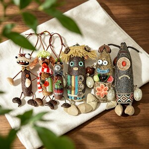 Puede incluir: Una colección de seis figuras caprichosas hechas a mano, cada una con características únicas. Están hechas de materiales naturales, con detalles como ojos de botón, detalles de tela y extremidades de alambre. Las figuras están dispuestas sobre una tela blanca.