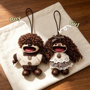 Puede incluir: Dos adornos de muñecas hechos a mano, uno etiquetado "Boy" y el otro "Girl". Las muñecas tienen cabello rizado castaño, bocas abiertas y visten trajes de punto. La muñeca del niño tiene un suéter con botones, y la muñeca de la niña tiene un vestido con un lazo.