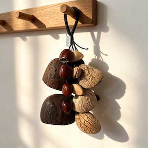 Puede incluir: Adorno colgante decorativo compuesto por elementos naturales. Incluye vainas texturizadas de color marrón oscuro, cuentas de color marrón rojizo y vainas de semillas claras y estampadas, suspendidas de un cordón negro. El adorno cuelga de un perchero de madera.