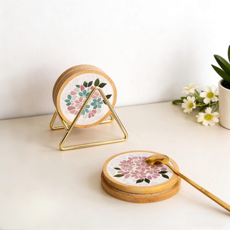 May include: A set of wooden coasters with a floral mosaic design in pink, blue, and green. The coasters are displayed in a gold metal holder. A gold spoon rests on top of a coaster. The coasters are a decorative home accessory.